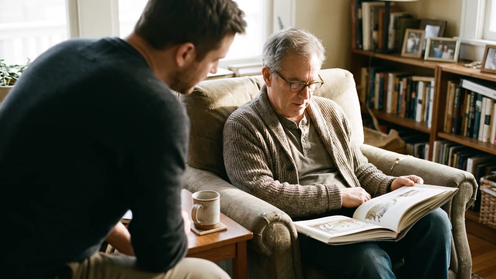 Un père en cardigan, installé dans un fauteuil, feuillette un livre illustré avec son fils adulte dans un salon